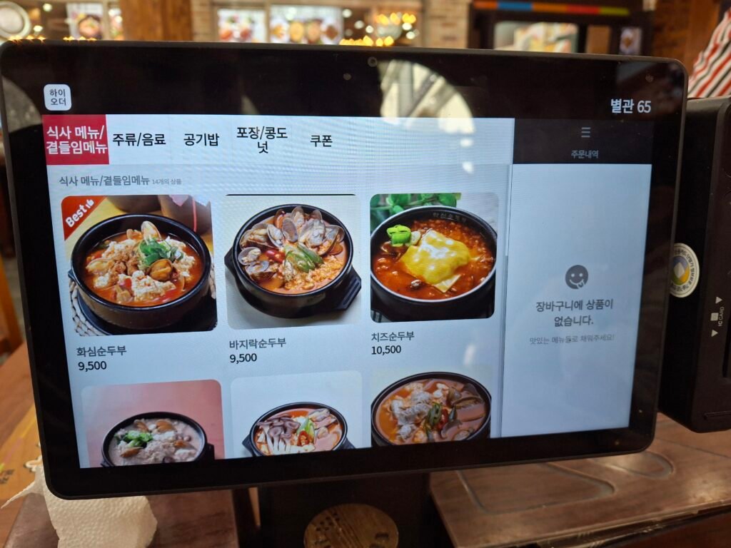 A modern smart ordering kiosk on a wooden table at a traditional Korean restaurant, allowing customers to easily customize their tofu stew.